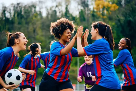 Women’s soccer team celebrating a goal during an outdoor match, showing teamwork, diversity, and sportsmanship.